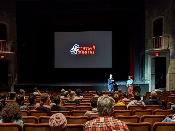 Two men standing before audience with Cornell Cinema logo behind them
