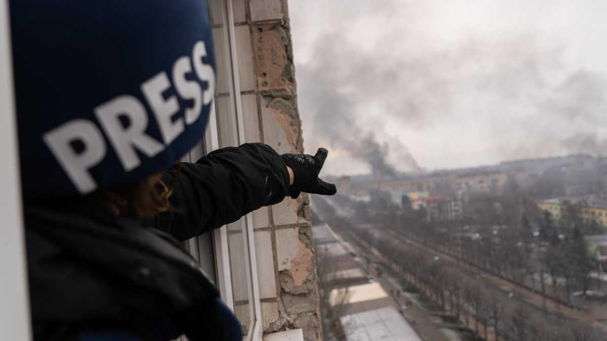 A close up photo of a blue "Press" helmet on a man pointing at a destroyed city skyline in the distance.