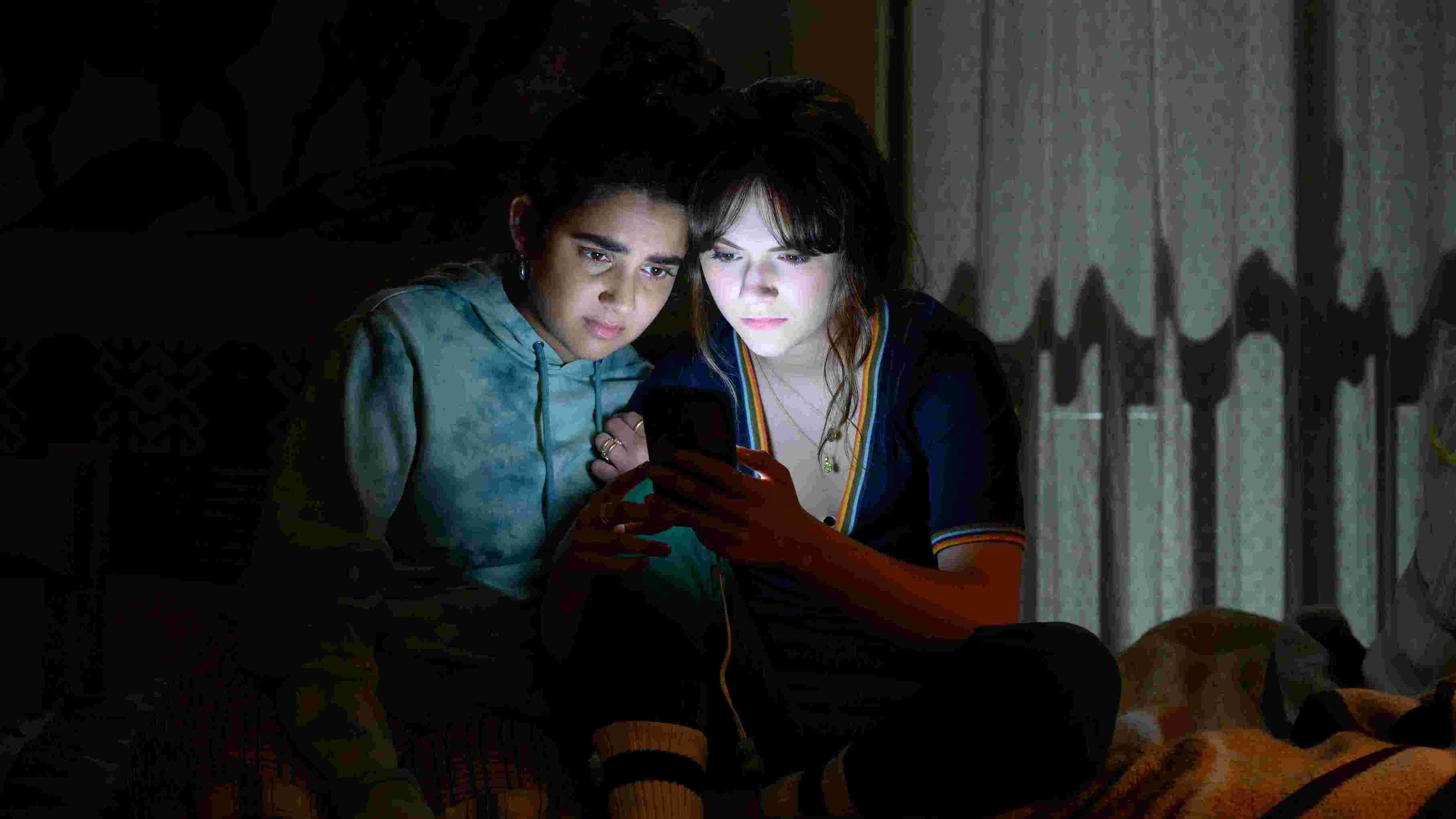 Two young women huddle in the darkness their faces illuminated by the light of a cell phone they are both looking at.