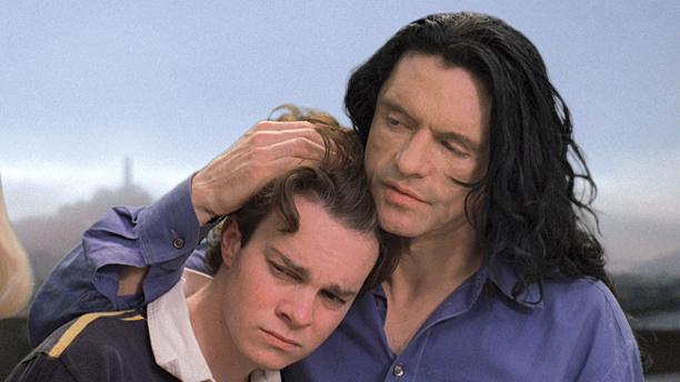 A young boy rests his head tenderly on the shoulder of a man with long dark hair who embraces him in return.