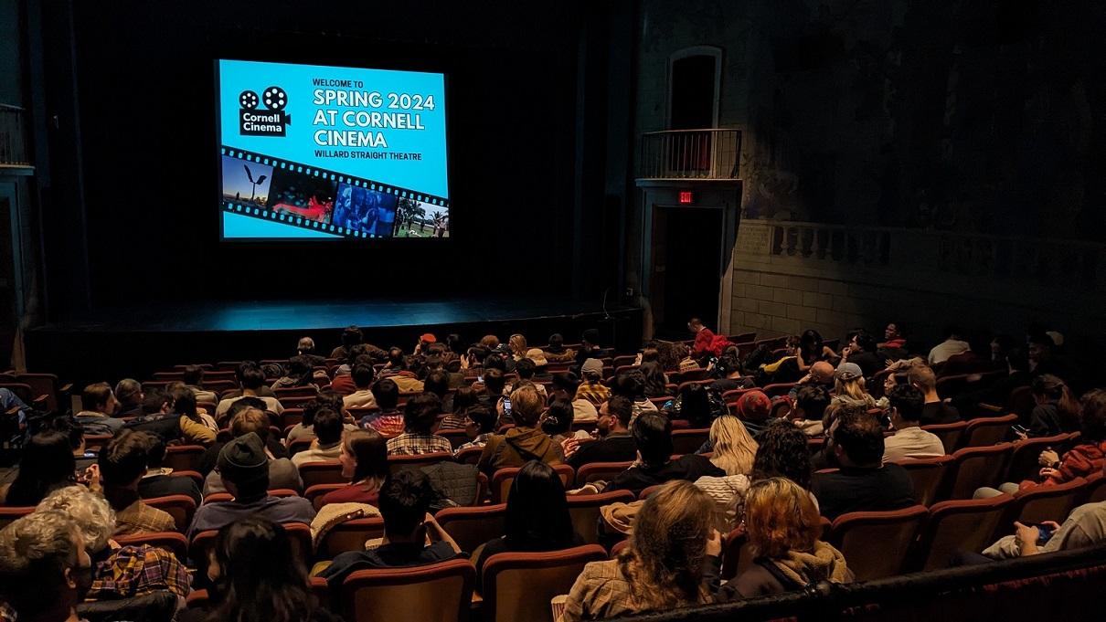 An old fashioned cinema will people seated facing a blue screen with black text.