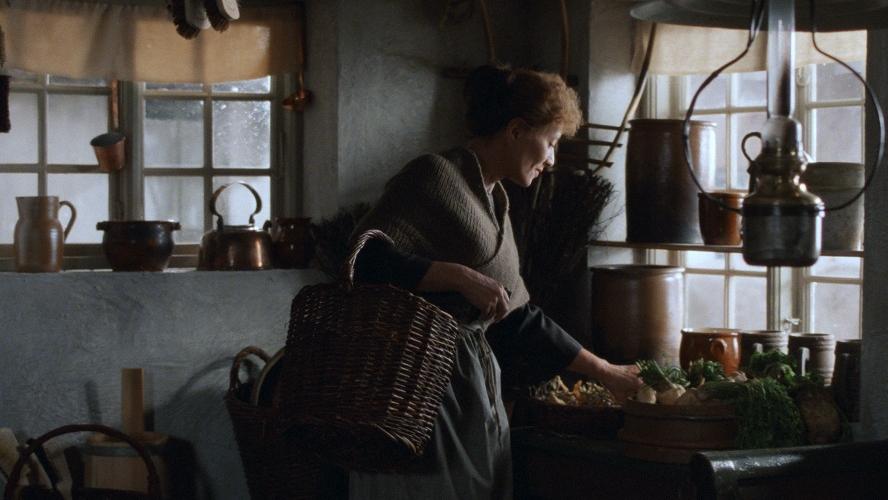 A woman holding a basket prepares food in a dimly lit kitchen.