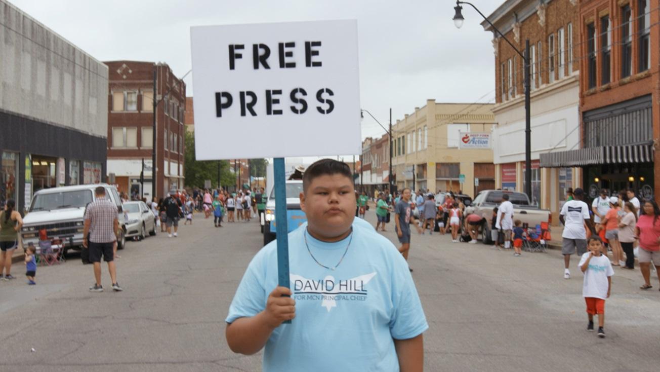 A young boy holding a sign that says "Free Press" and standing in the middle of a bustling street 