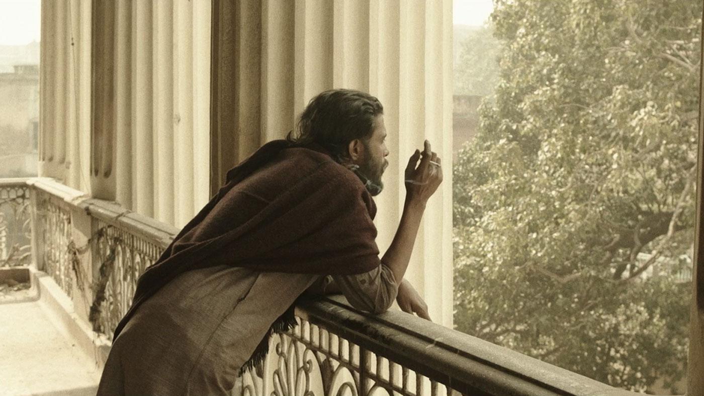 A man leaning against a balcony railing and smoking a cigarette overlaid with red and white text.