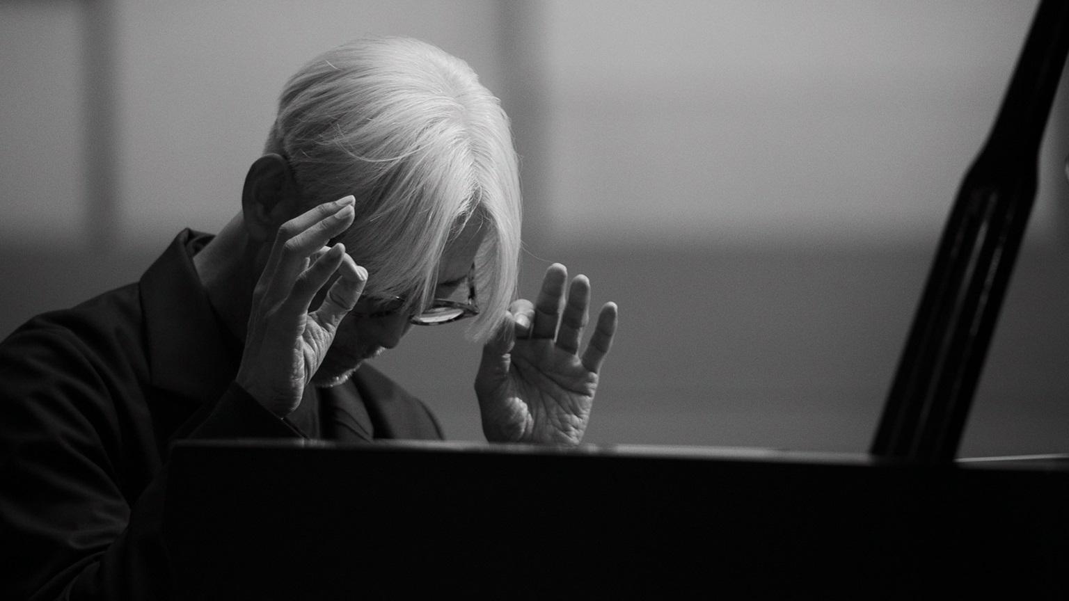 A black and white photo of a man gesturing while sitting at a piano.