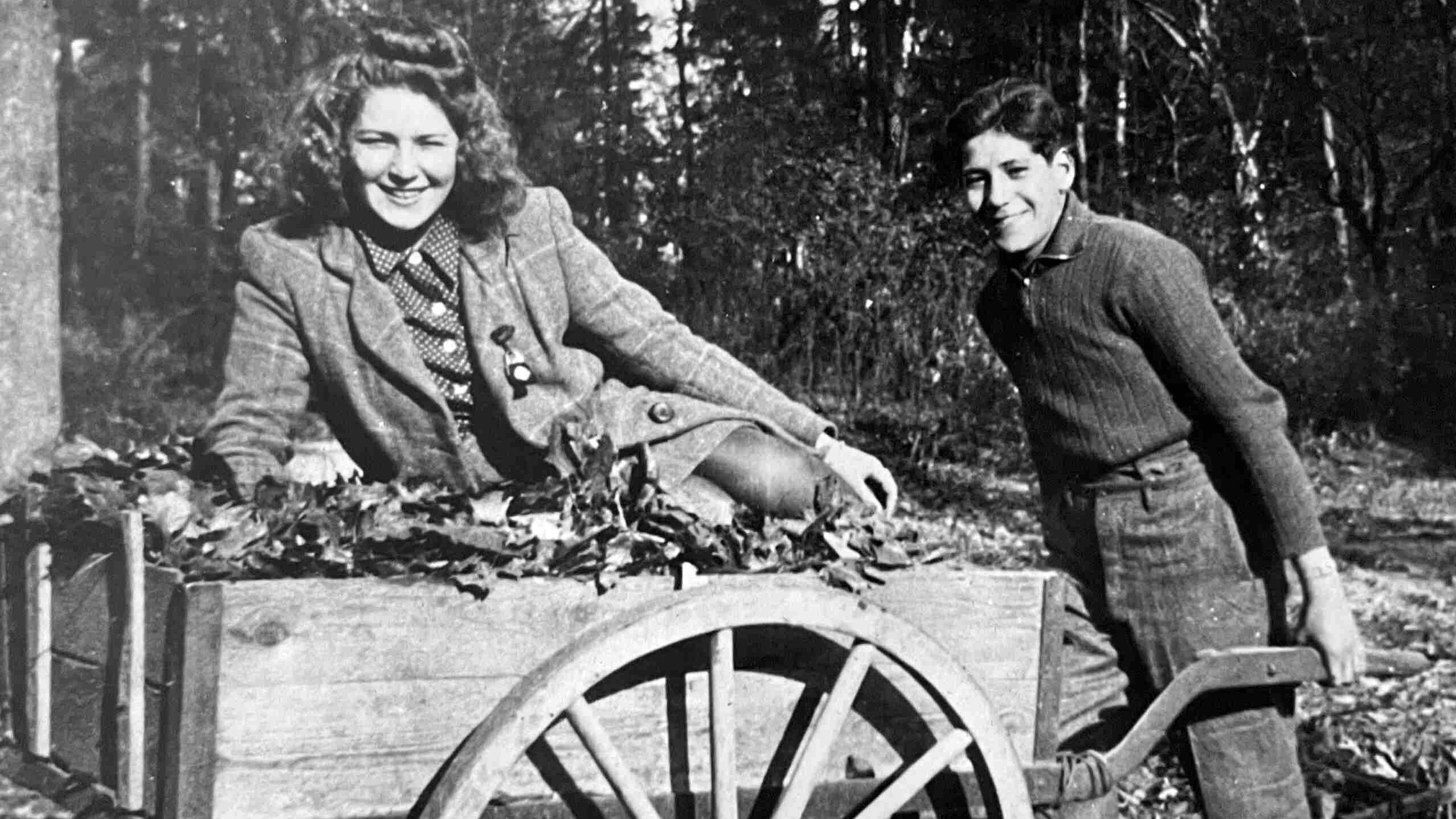 A black-and-white photograph of a yougng woman sitting in a wheel barrow being pushed by a young man.