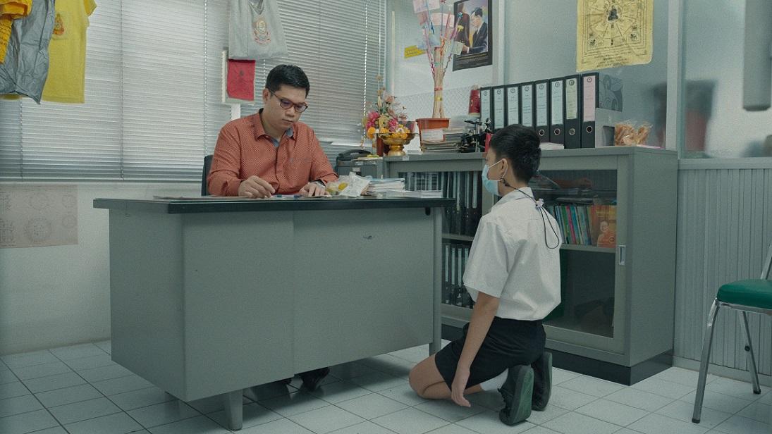 A boy kneeling in front of a desk where a teacher is working