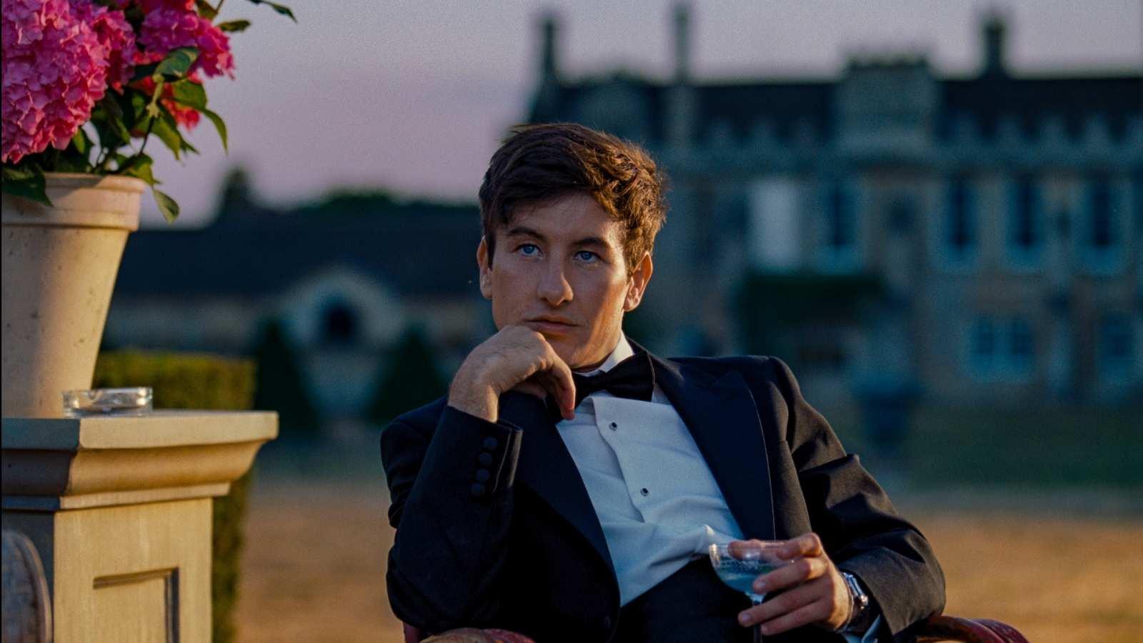 A man in a tuxedo holding a glass of champagne while sitting next to a flower pot with a grand home in the distance.