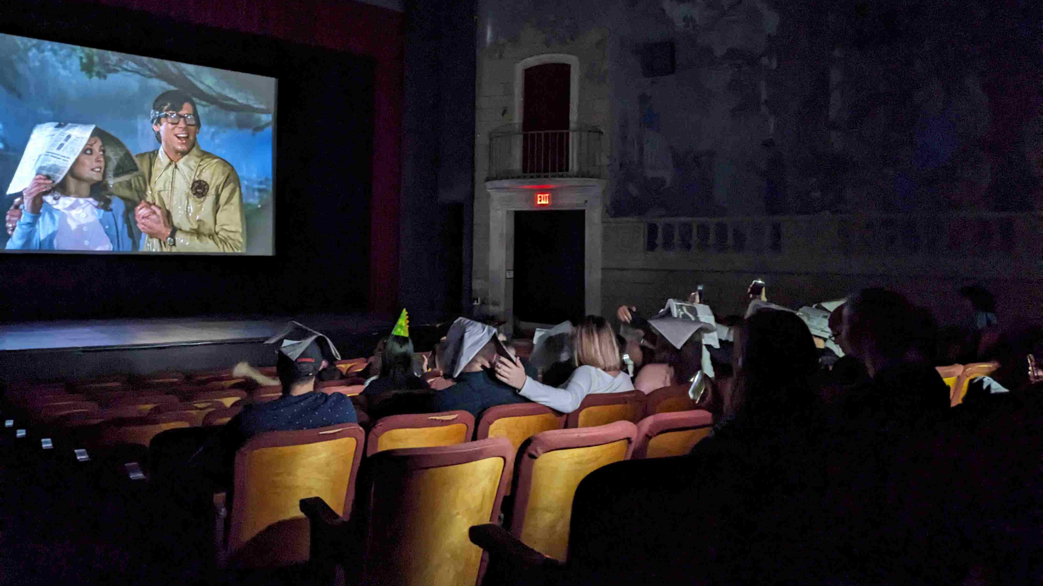 A group of people watching a movie wearing party hats and newspapers on their heads.