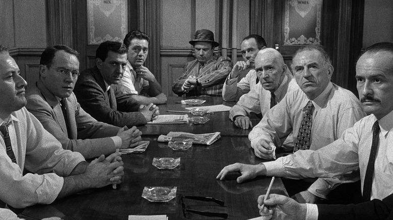 A black and white photograph of eleven individuals sitting around a long table. The setting appears to be a formal meeting or discussion, with most individuals wearing suits and ties. Several documents or papers are visible on the table in front of them.