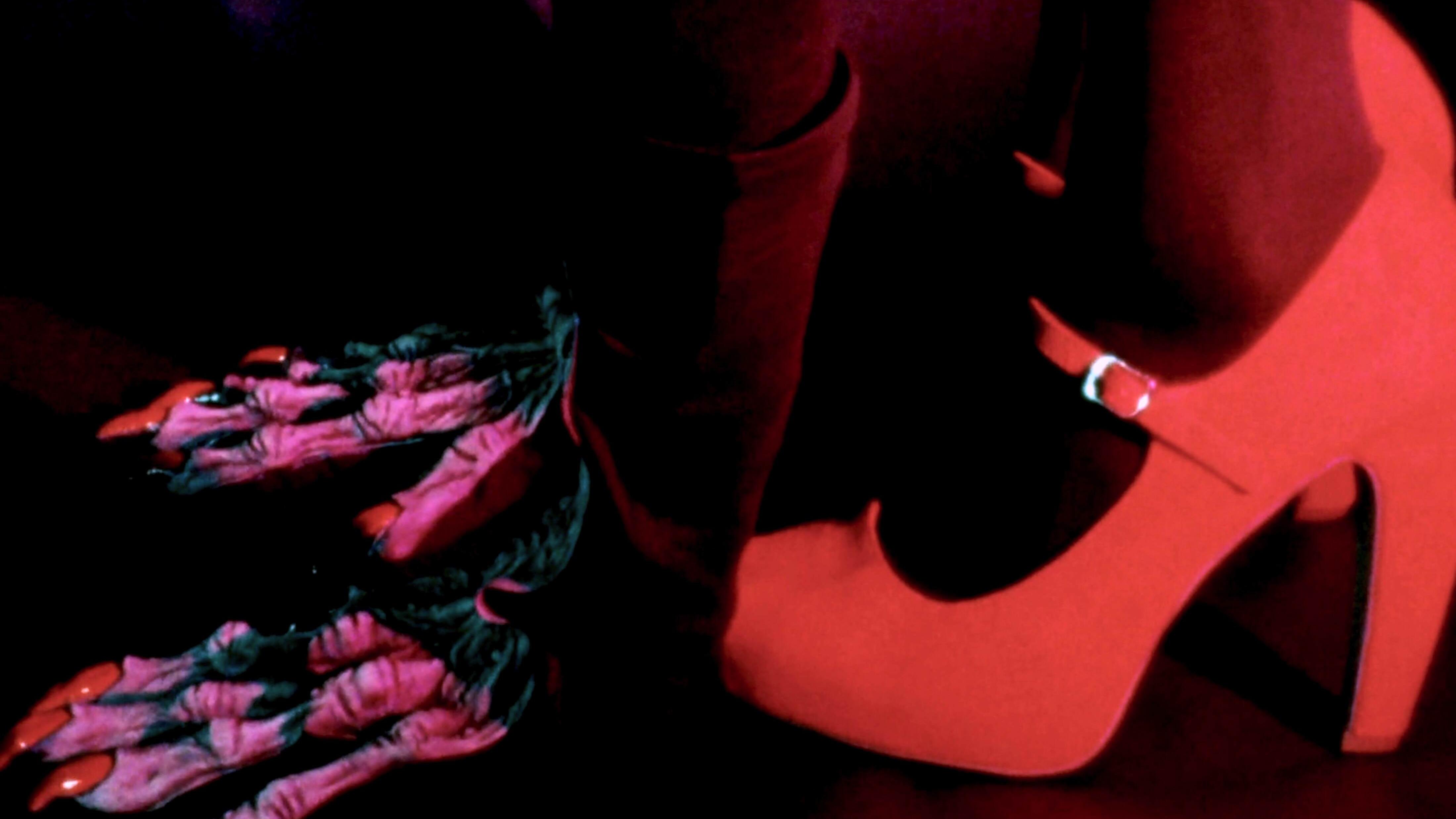 Close-up of a person’s lower legs and feet. One foot is wearing a high-heeled shoe, while the other foot is bare and stepping on a piece of clothing or fabric on the floor. The lighting has a reddish-purple hue, adding a dramatic effect to the scene.