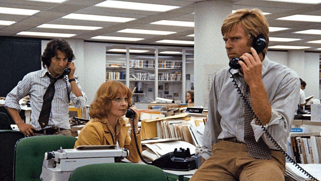 An office scene from a past era with three individuals working amidst typewriters and telephones. Desks are cluttered with papers and office equipment, suggesting a busy work environment.