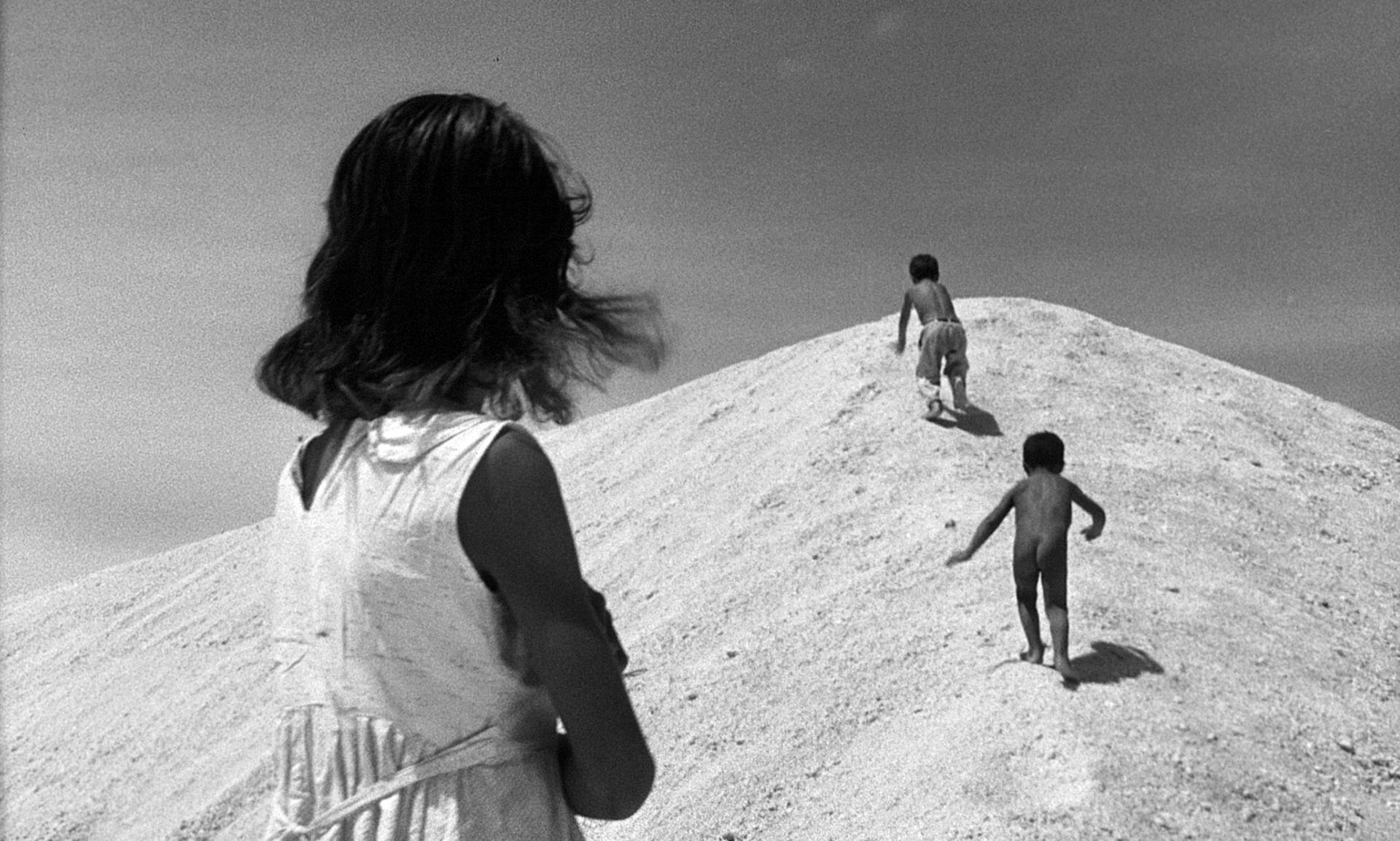 Black and white photograph of three individuals, likely children, on a mound of earth or sand. The foreground shows the back of one individual with long hair, looking towards the other two who are further up the mound. The sky is clear and bright, suggest