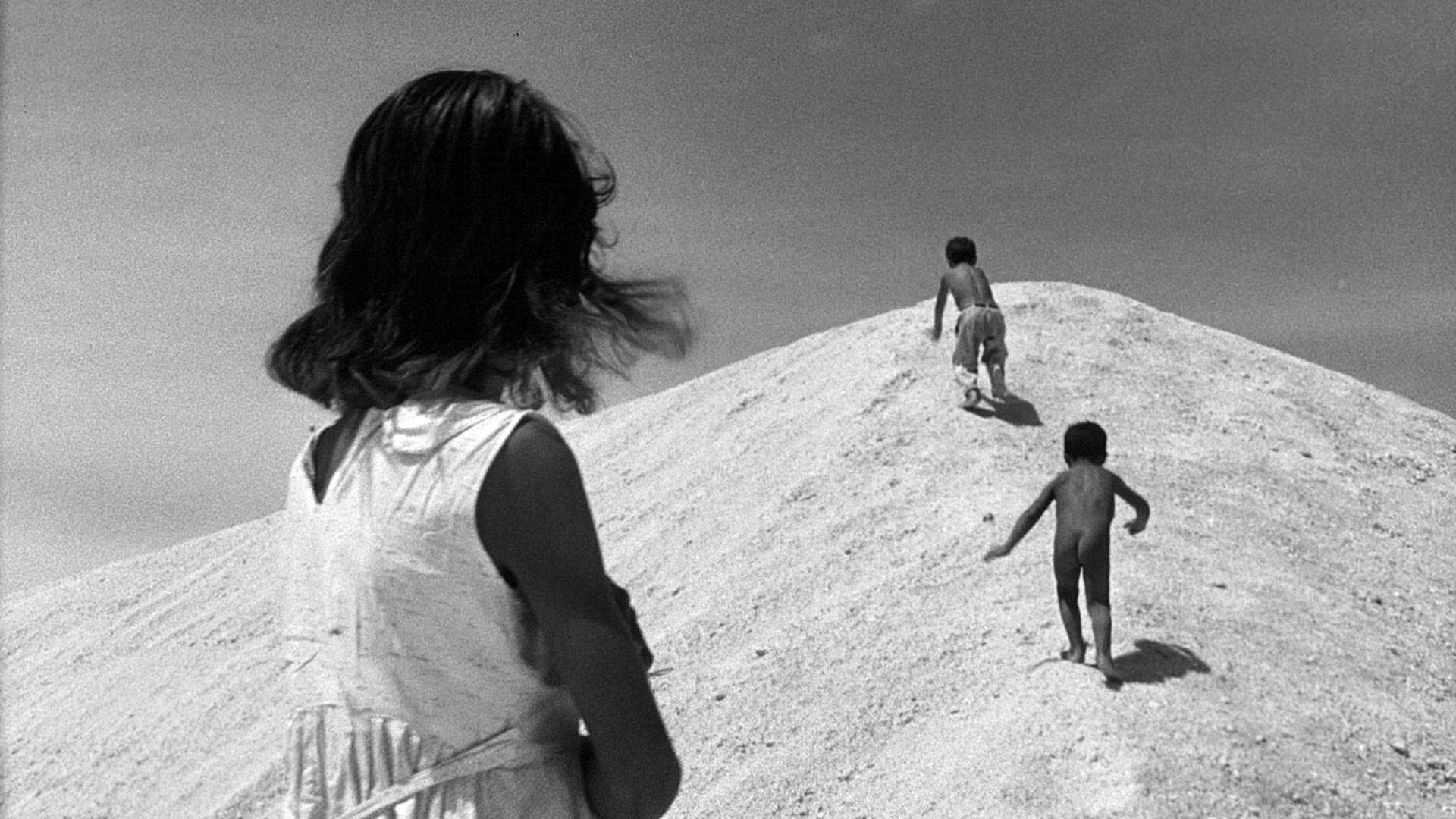 A black and white photo of two children climbing a pile of sand with an older child looking on.