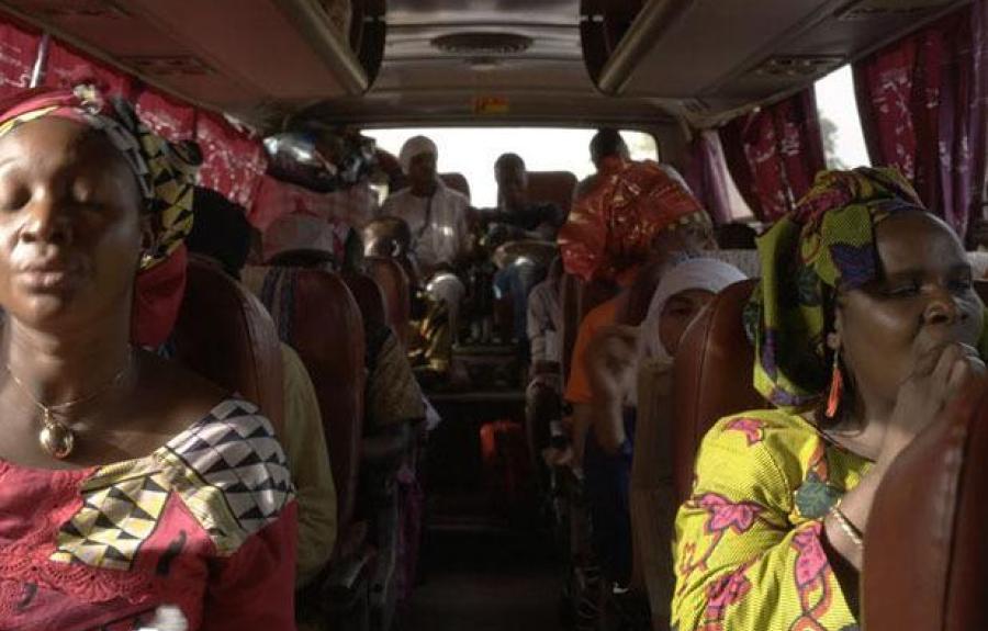 Interior of a crowded bus with passengers seated facing forward. The bus appears to be in motion, and the passengers are dressed in various colorful garments. Luggage is stored above the seats, and the windows are covered with curtains. The atmosphere see