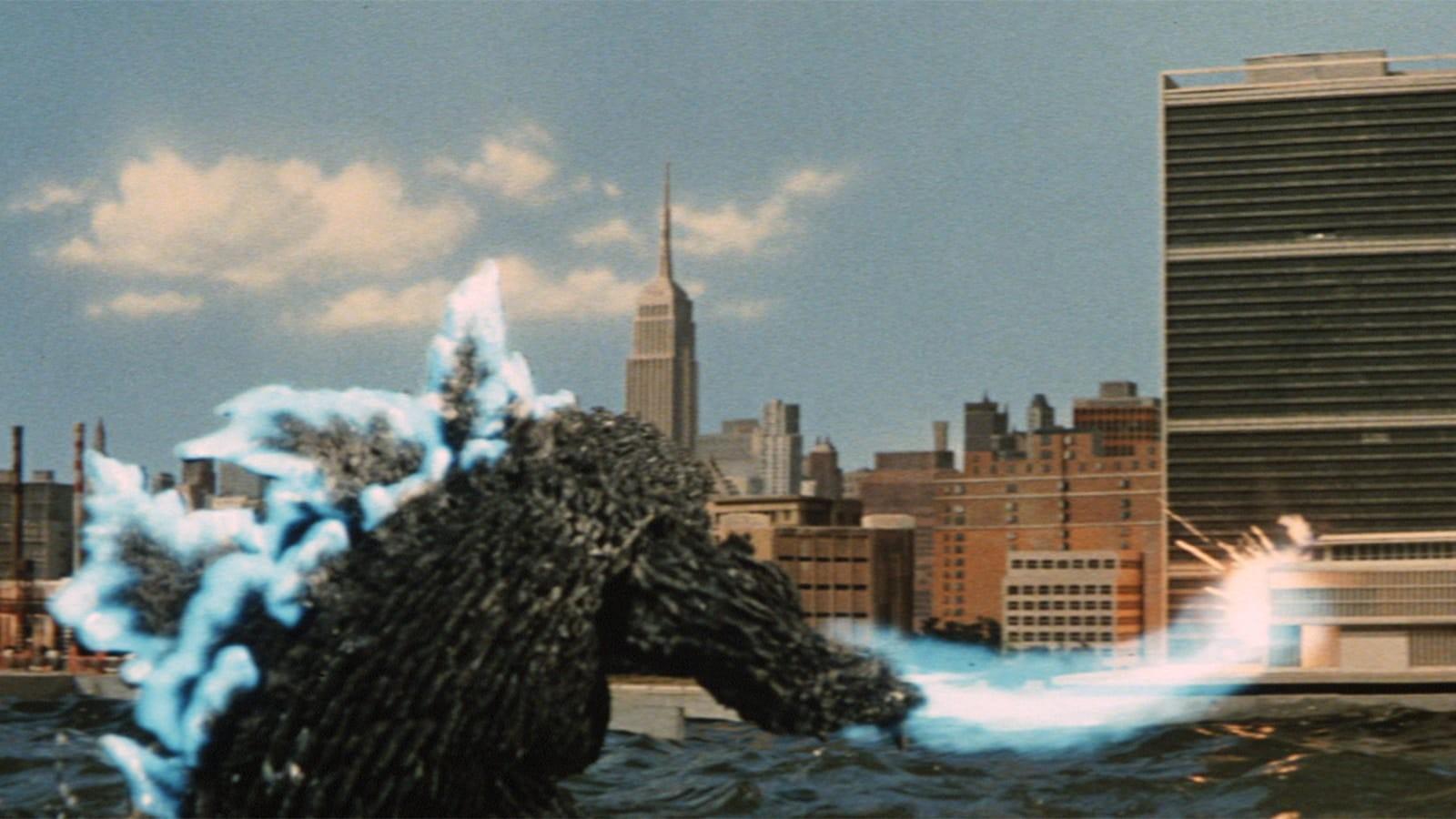 A large, dark, reptilian creature resembling Godzilla standing in a body of water with a city skyline in the background. The creature is emitting a blue beam from its mouth towards the right side of the image. The skyline features several high-rise buildi