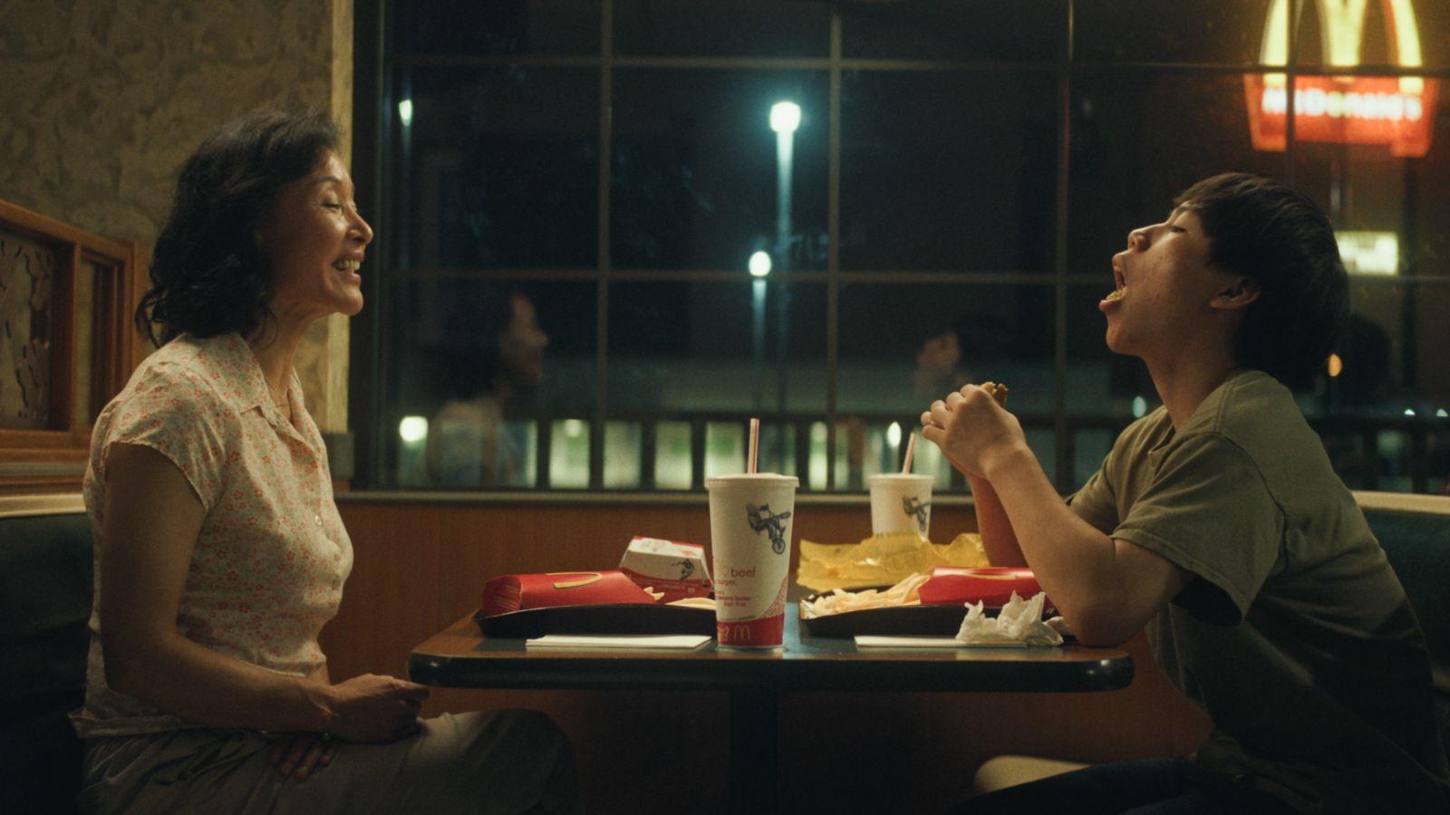 Two individuals seated opposite each other at a table inside a fast-food restaurant at night. On the table, there are various fast-food items including a drink with a straw, food wrappers, and partially eaten meals. The bright signage of McDonald’s is vis