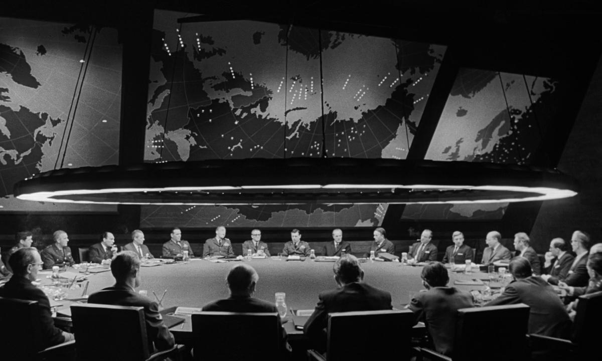 A black and white photo of a group of men seated around a large, oval-shaped table in a room with dim lighting. Above the table hangs a circular light fixture that illuminates the area directly below. Behind the men is an expansive wall map displaying var