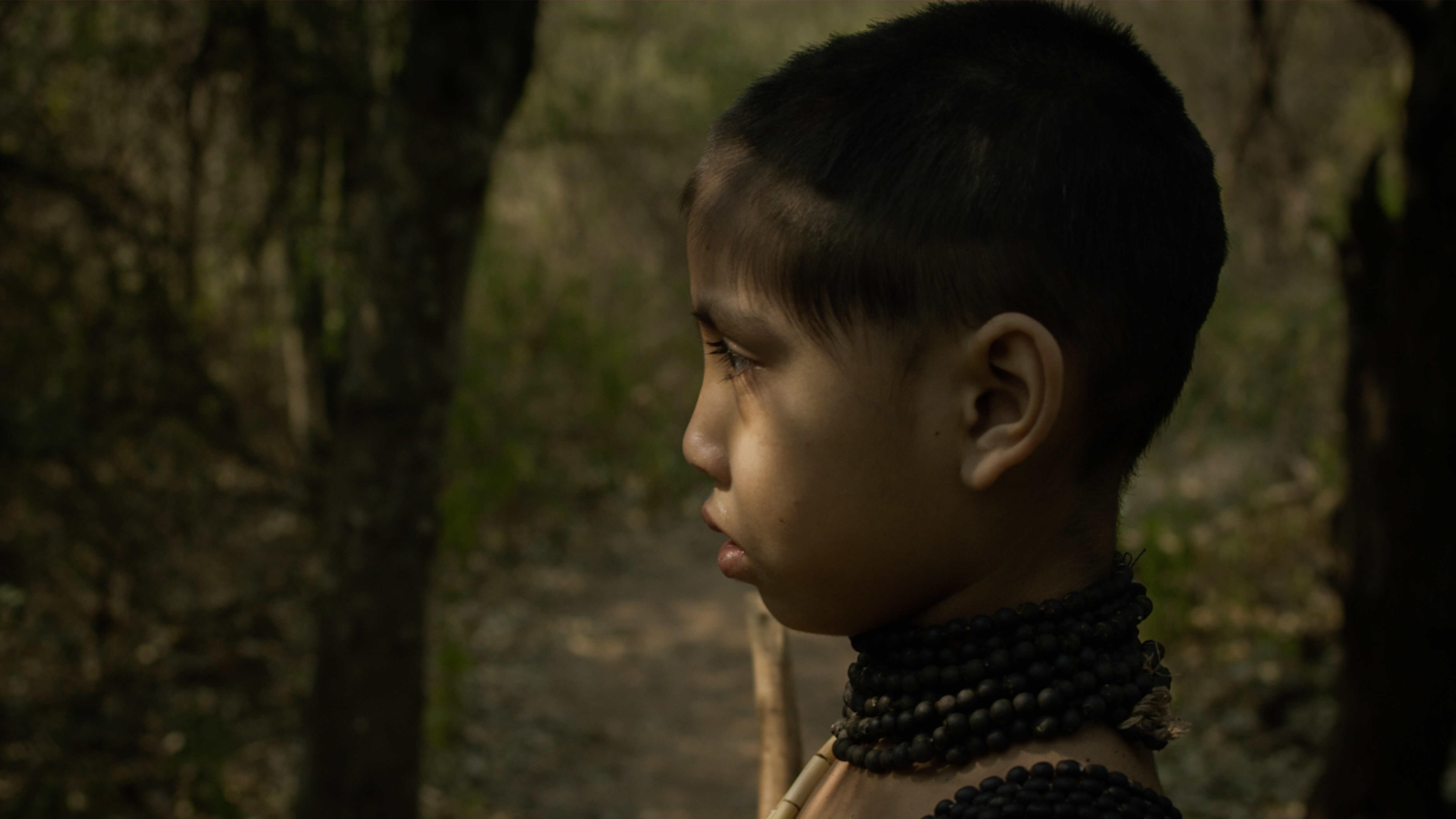 A close up of a young child looking into the distance while wearing a black beaded necklace with sunlight dabbling their face.