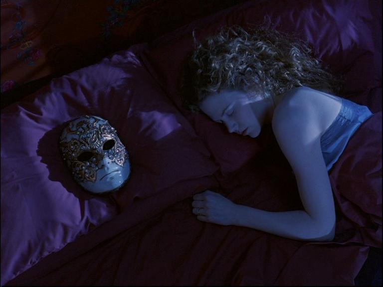 A person with light-colored hair is lying on a bed with purple bedding, appearing to be asleep. Next to the person is an ornate mask with gold detailing and white feathers on a dark background. The scene has a nocturnal ambiance, suggesting it could be ni