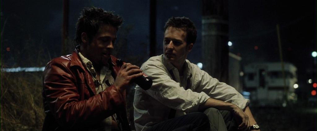 Two individuals are sitting side by side at night. The person on the left is wearing a red jacket and holding a bottle, while the person on the right is dressed in a white shirt. They are seated in an outdoor setting with industrial elements, such as a la