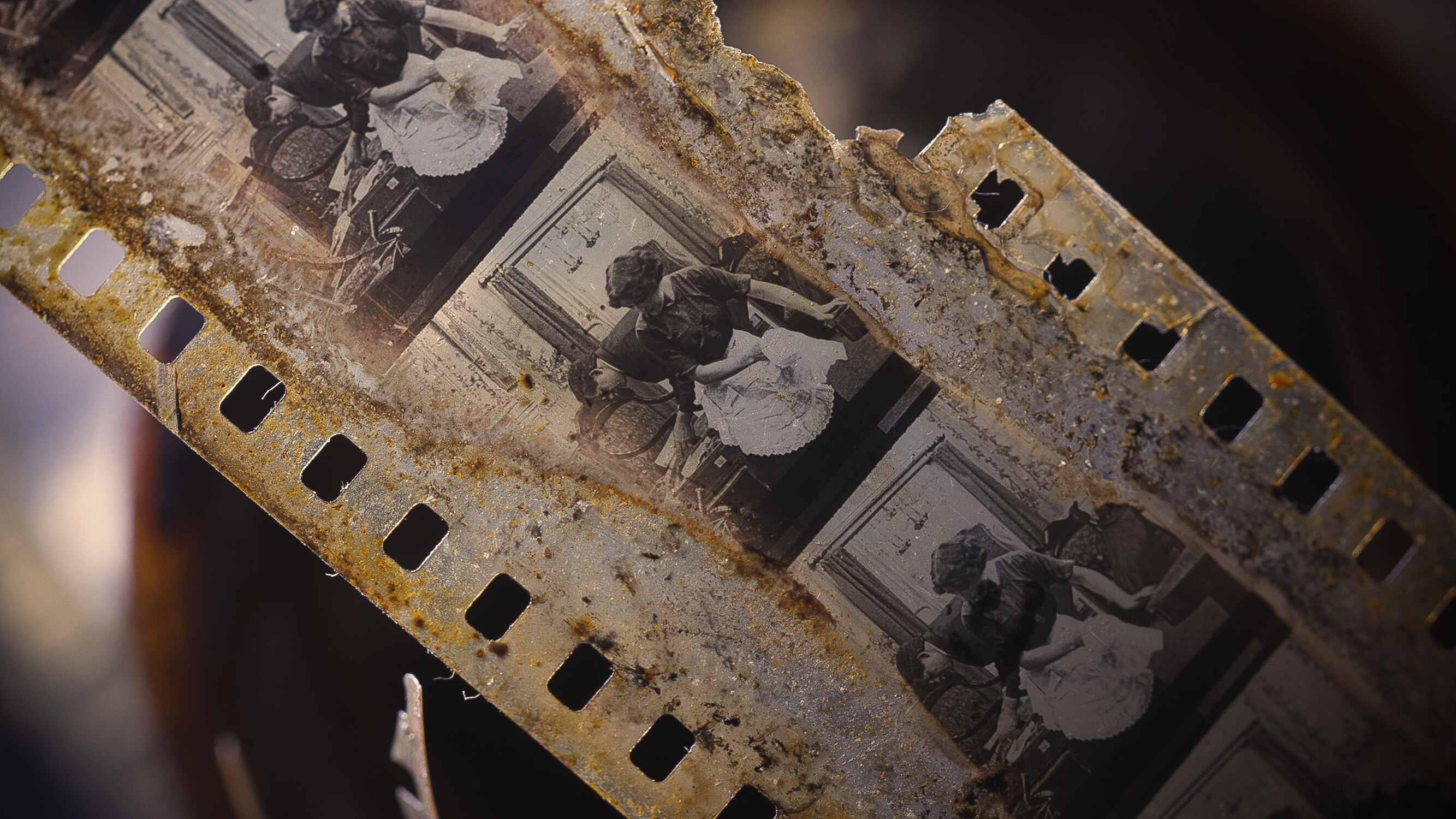 An old, deteriorated film strip with a series of black and white images. The film strip has visible damage and rust, indicating age and decay. The images on the film appear to be from an early 20th-century performance or play, featuring individuals in per