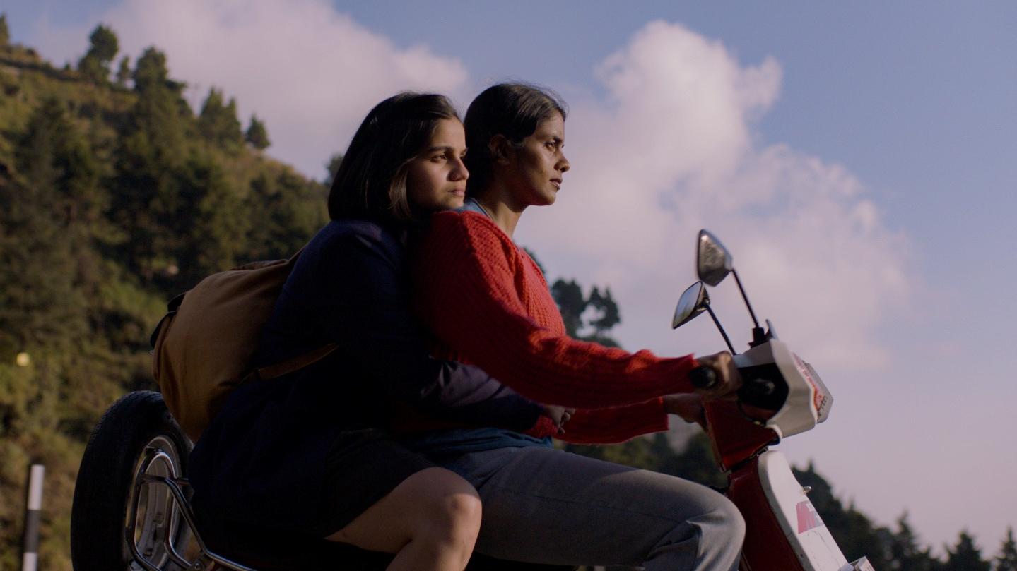 A close-up image of mother and daughter riding a moped against a cloudy blue sky.