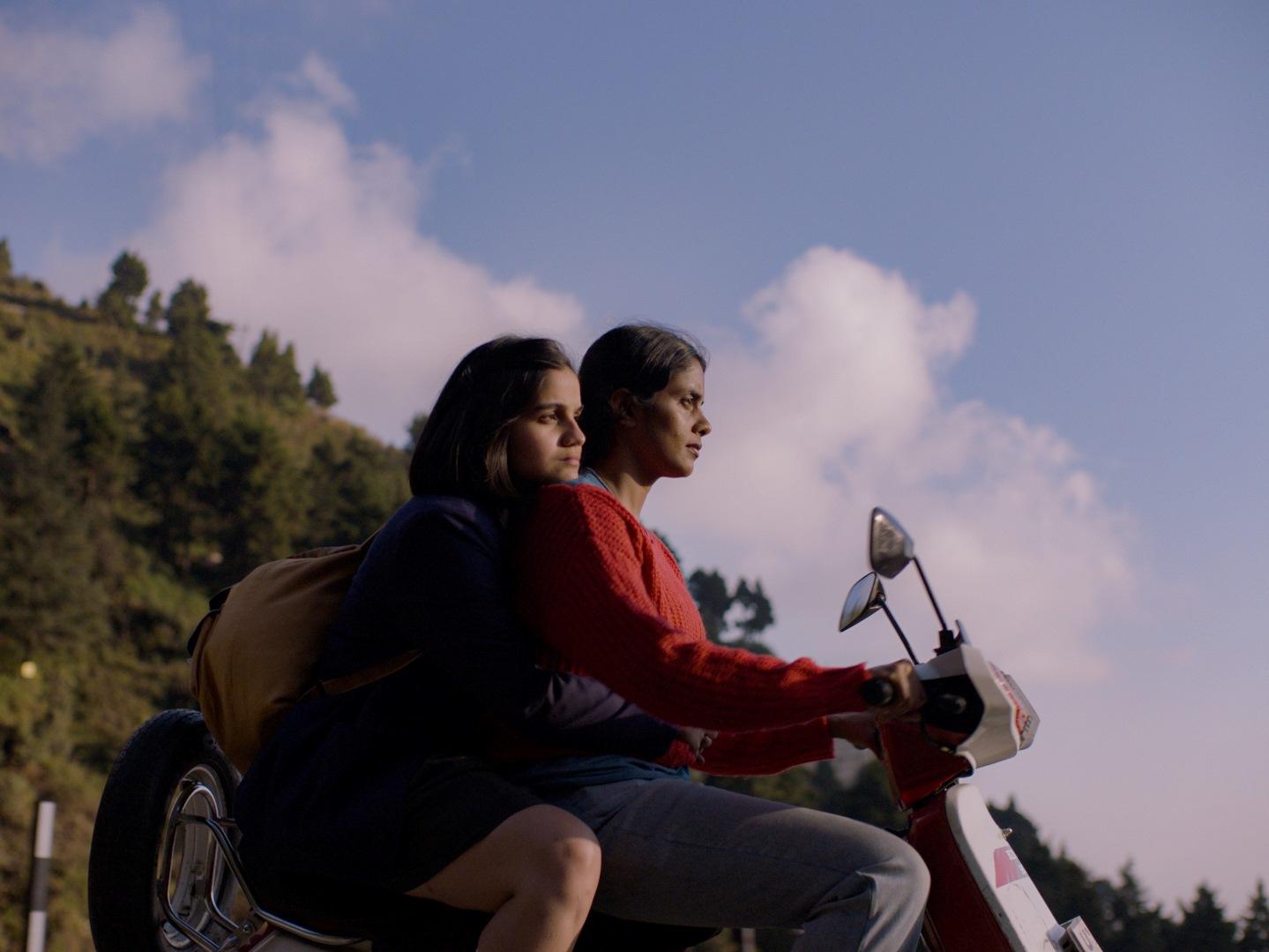 Two individuals on a scooter are stationary with a backdrop of a hill covered in greenery under a clear sky. The person in the front appears to be controlling the scooter while the one at the back is sitting close, possibly holding onto the driver or the 