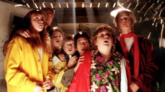 A group of children dress in colorful clothing standing in a basement beneath wooden floorboards.