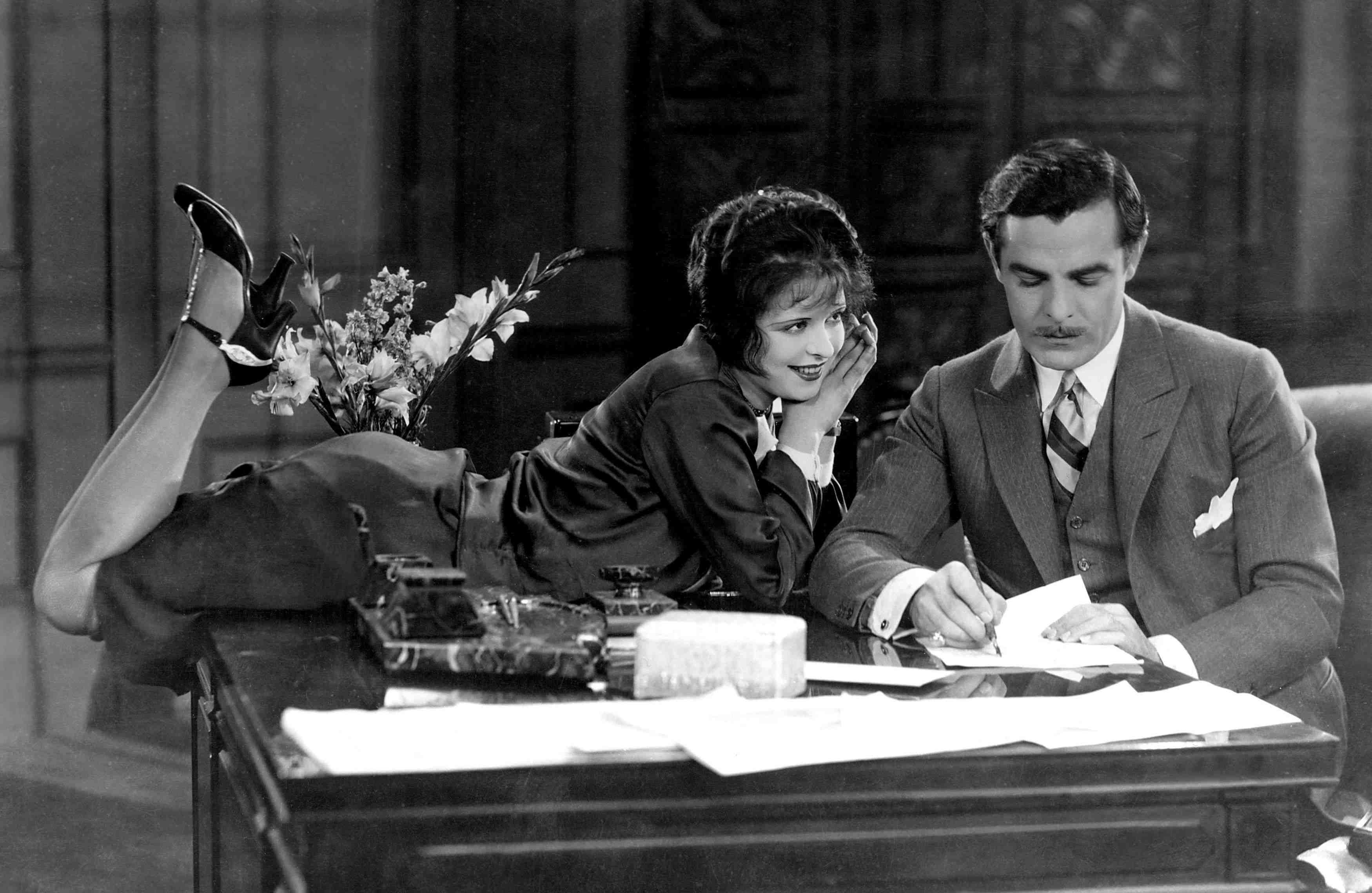 A black and white photograph featuring two individuals sitting at a desk. One individual appears to be reclining with legs crossed on the desk, while the other is seated upright, seemingly writing or signing a document. On the desk, there are various item