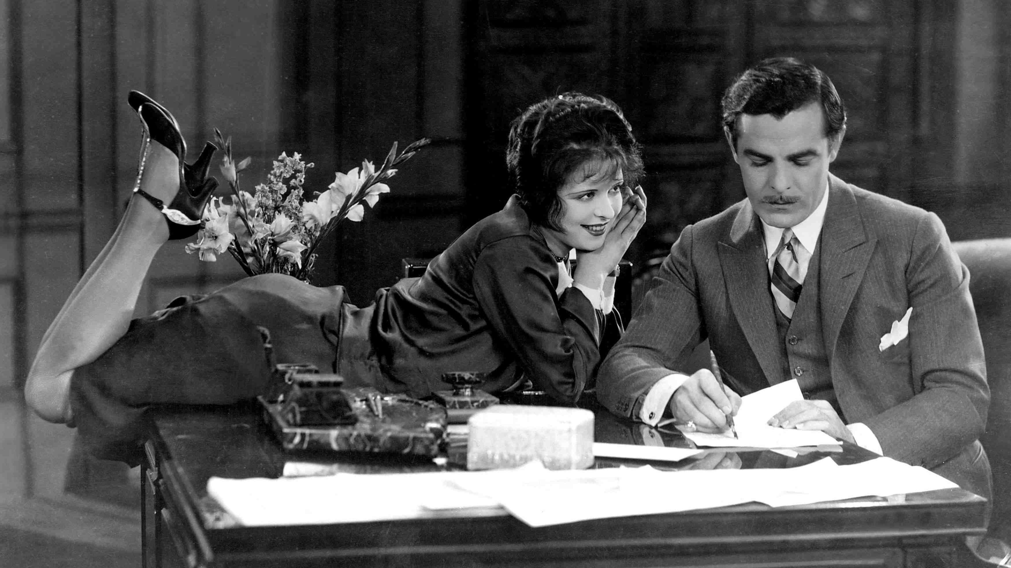 An old fashioned photograph of a women perched on a desk looking longing at a man who is ignoring her and writing on a piece of paper.