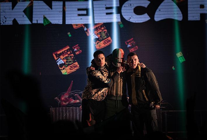 Three individuals on a stage. Behind them, there is a large screen displaying the word ‘KNEECAP’ in bold capital letters, and below it are images of what appear to be currency notes. The setting suggests that this is a live performance or event, and the w