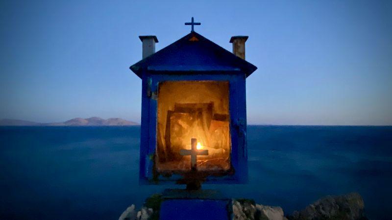 A small shrine or religious icon display with a Christian cross on top. It is set against a blurred background featuring what appears to be a body of water and possibly distant land or mountains. The shrine is illuminated from within, casting a warm glow,