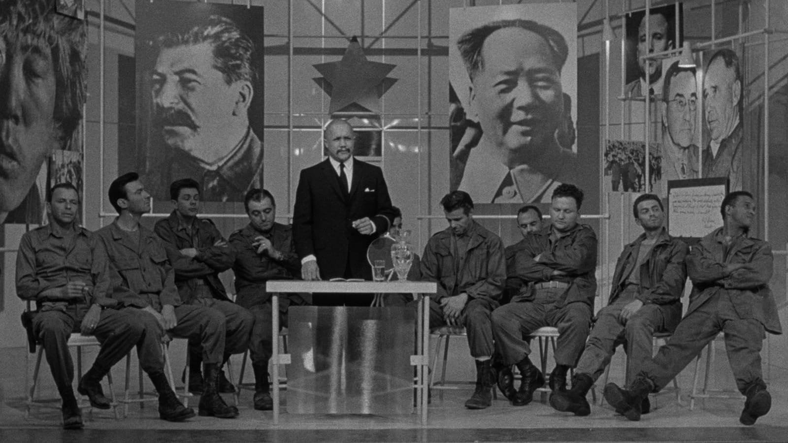 A black and white photograph featuring multiple individuals seated in a row. Standing behind the seated individuals is another figure, also with their face obscured, positioned next to a table with what appears to be a microphone on it. The background inc