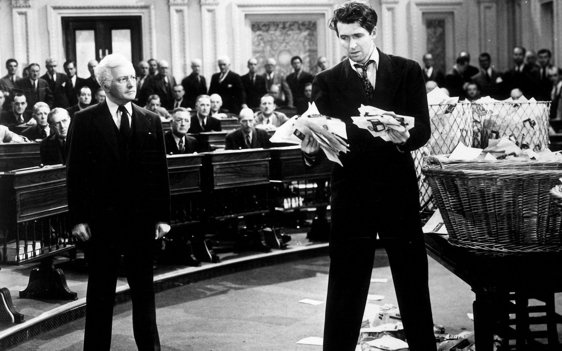 A black and white photograph showing a scene with multiple individuals in what appears to be a legislative assembly or courtroom. One of these individuals is standing at a podium, holding papers, and appears to be addressing the assembly, while the other 