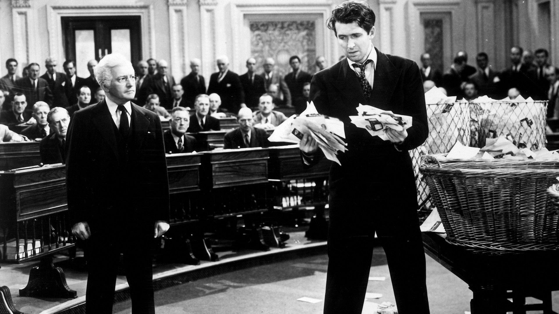 A forlorn looking man standing in the center of a courtroom holding a disorganized stack of papers in his hand.