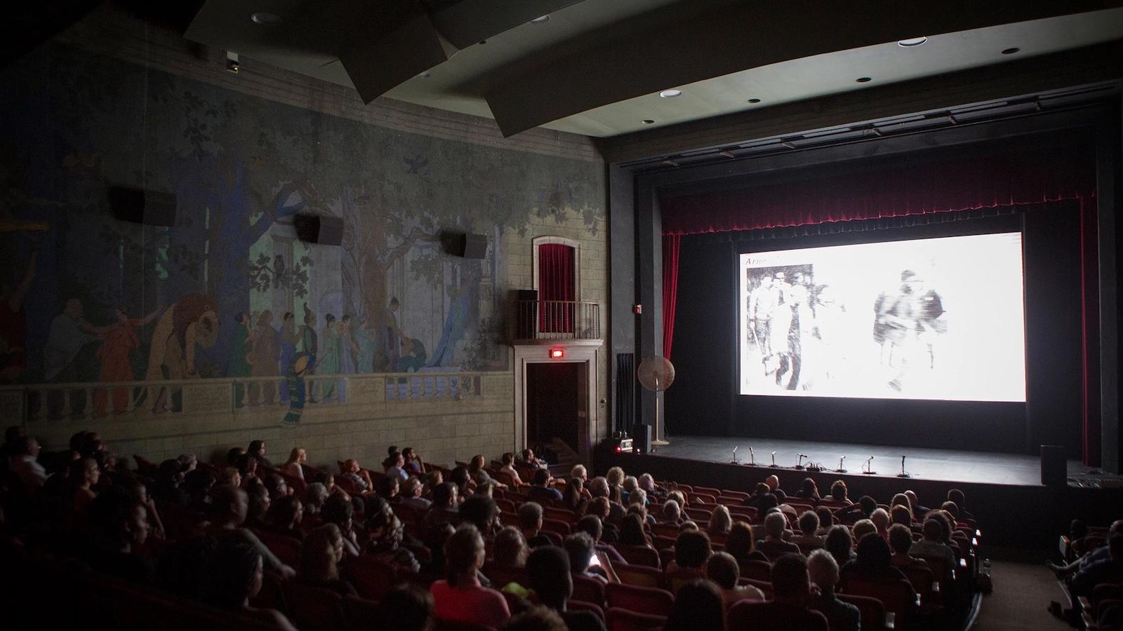 A crowded historic movie theater with an obscure black and white image on screen.