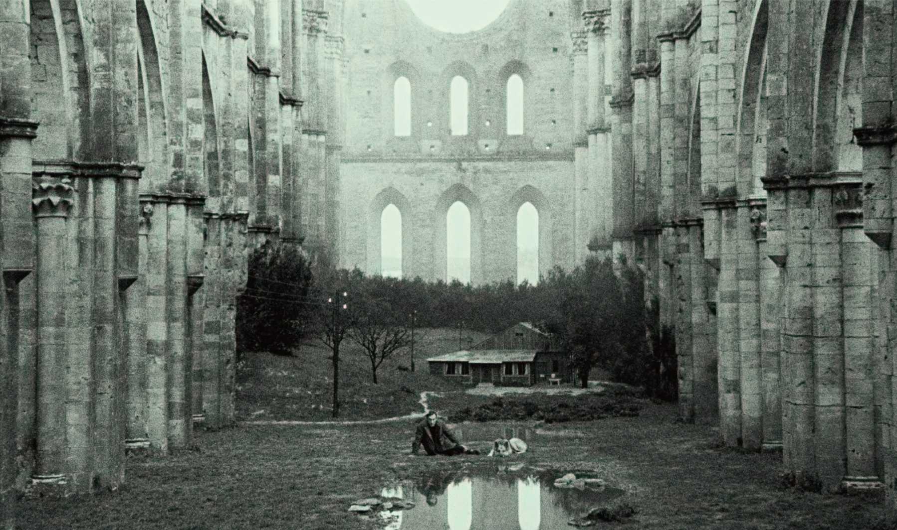 The ruins of a large gothic structure with tall arches and no roof. In the foreground, there is a small body of water reflecting part of the structure. A person is seated near this water, and there are several other individuals grouped together slightly f