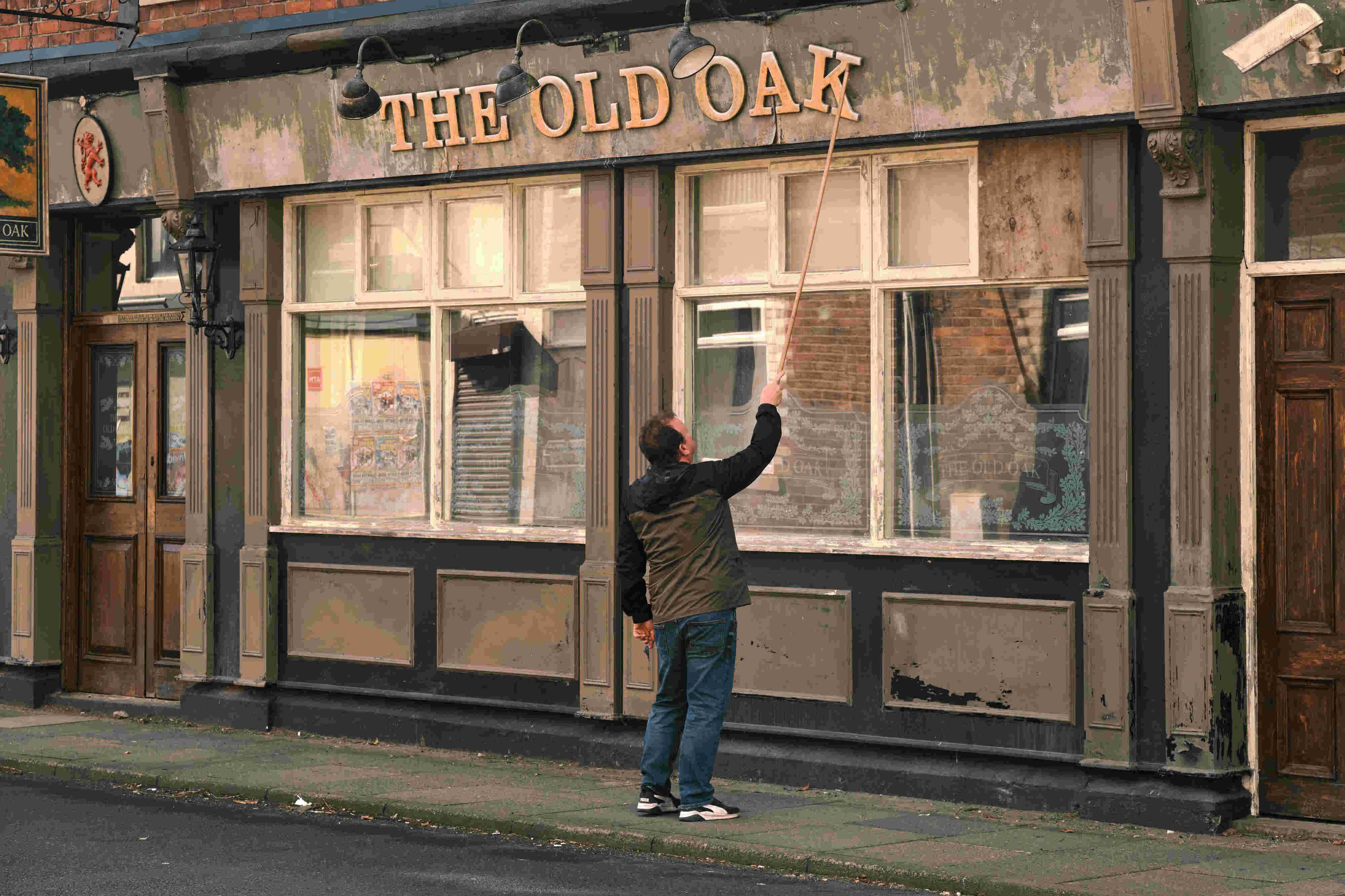 An individual standing in front of a building with a sign that reads “THE OLD OAK.” The person appears to be reaching up towards one of the crooked sign letters with a stick or tool to fix it. The building has a traditional facade, suggesting it might be 