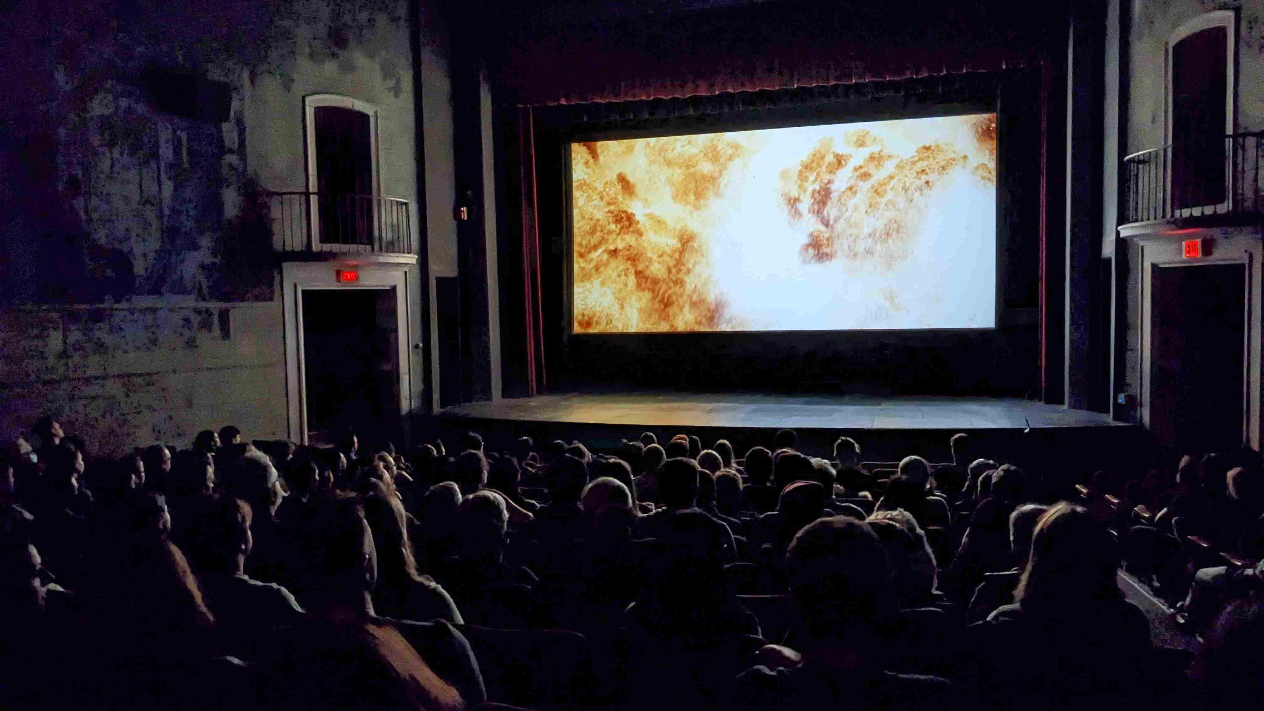 An audience sits in the dark looking at a movie screen filled with a fiery image from Christopher Nolan's Oppenheimer (2023).