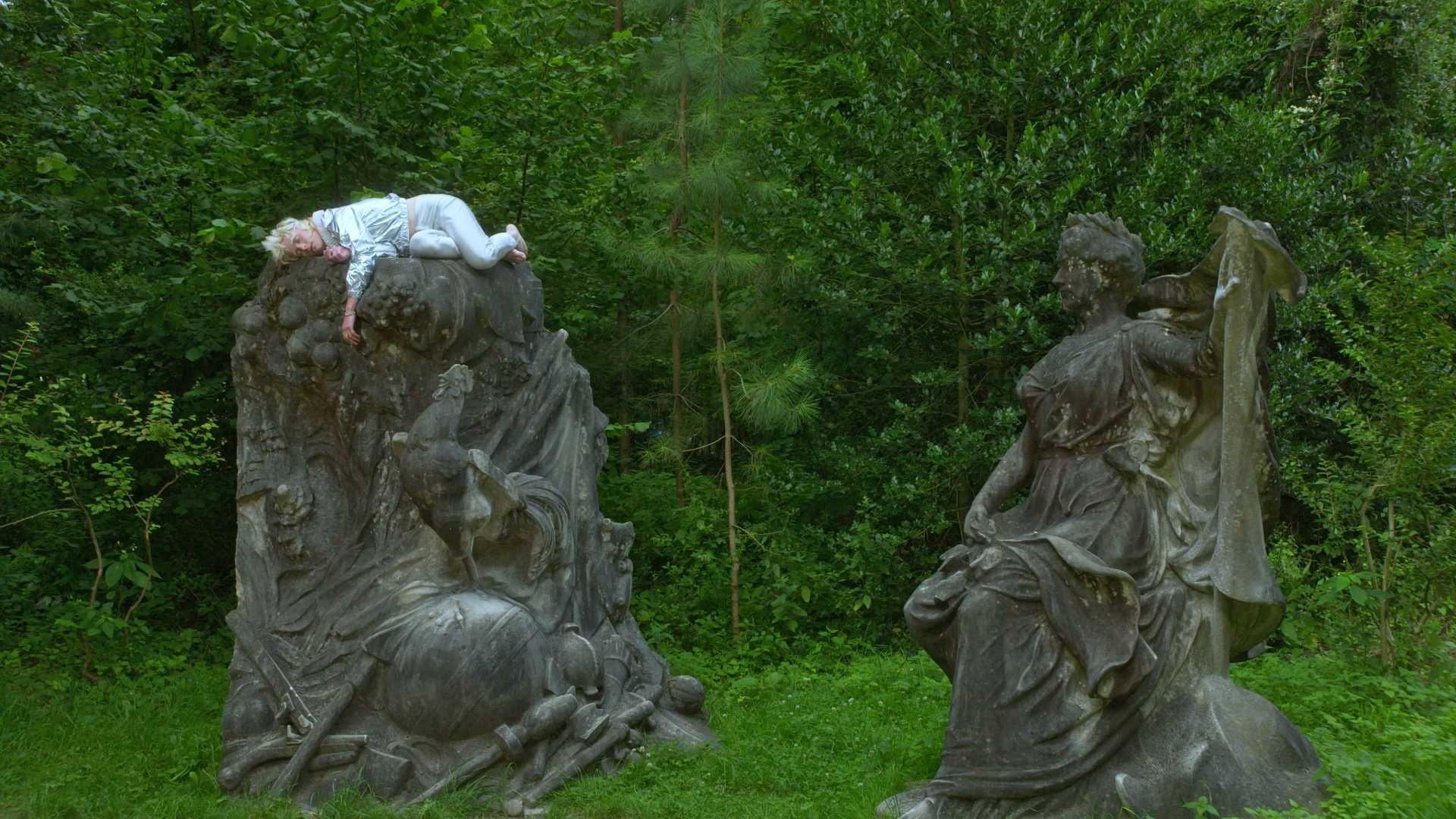 A man dressed in light blue sleeping atop a stone statue in a lush garden.