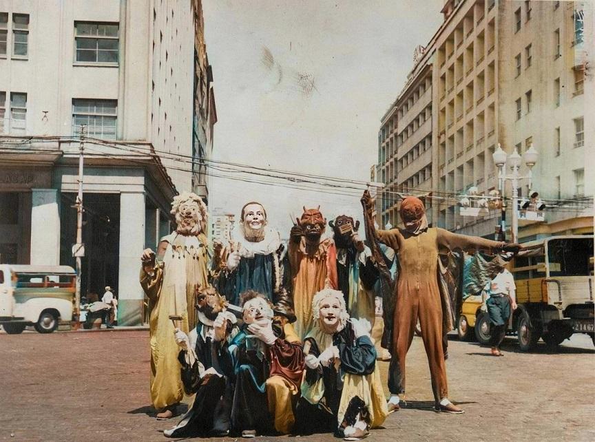 A vintage photograph of a group of people wearing carnivalesque costumes and masks while standing in the middle of an urban street.