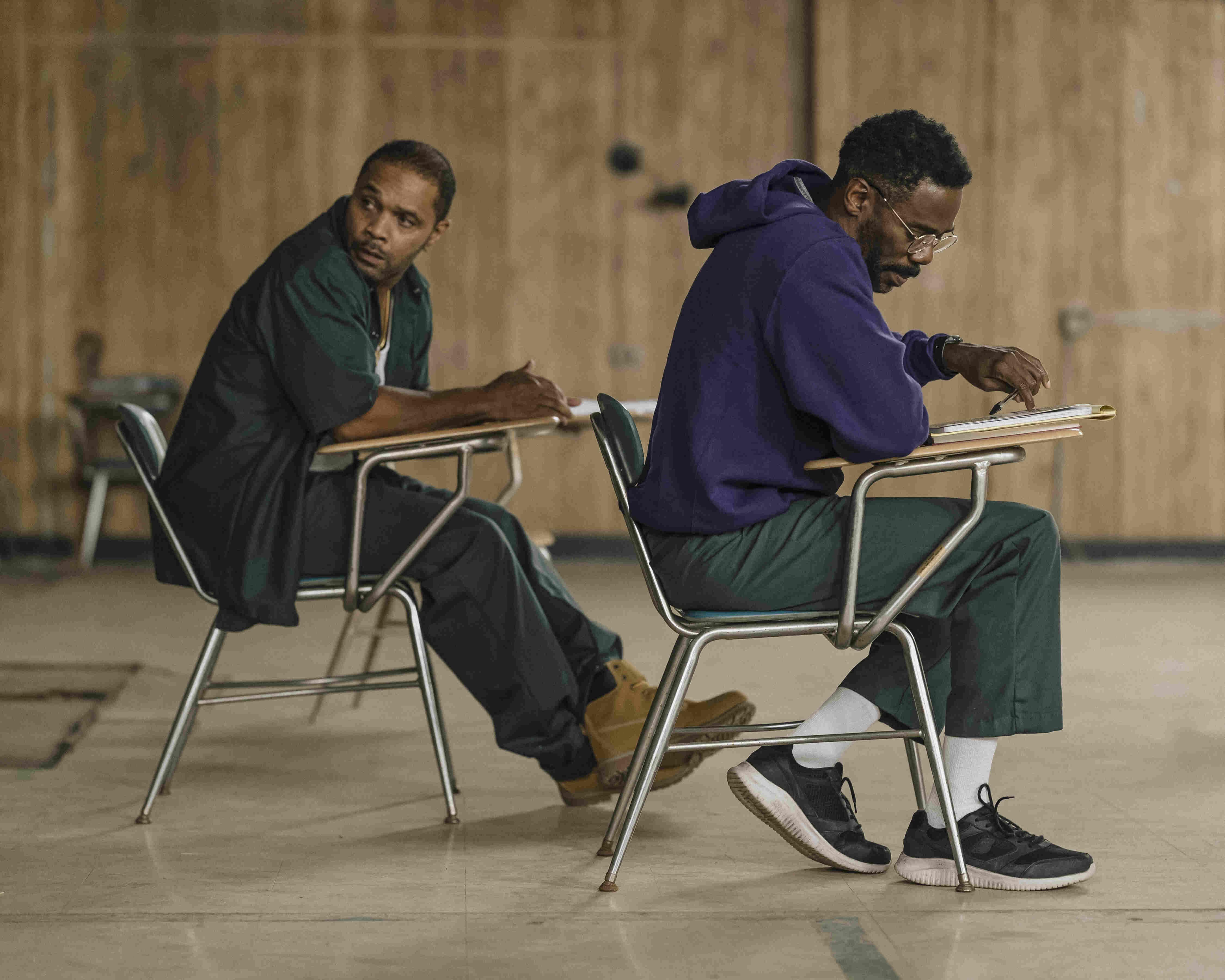 Two individuals seated on chairs at separate desks in a room with wooden paneling on the walls. Both individuals are wearing green pants and one is wearing a purple sweatshirt. The individual in the purple sweatshirt appears to be writing or drawing on a 