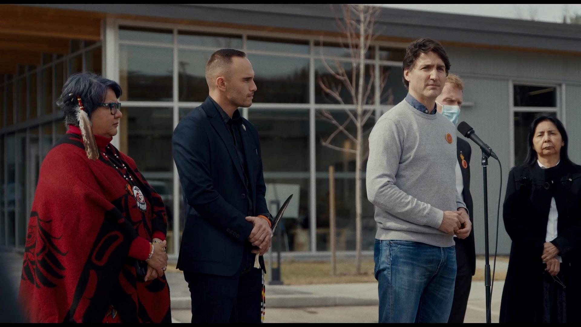 A group of five individuals standing outdoors. They appear to be gathered for an event or announcement, as indicated by the presence of a microphone in front of one individual. The setting includes a building in the background and it seems to be a cloudy 