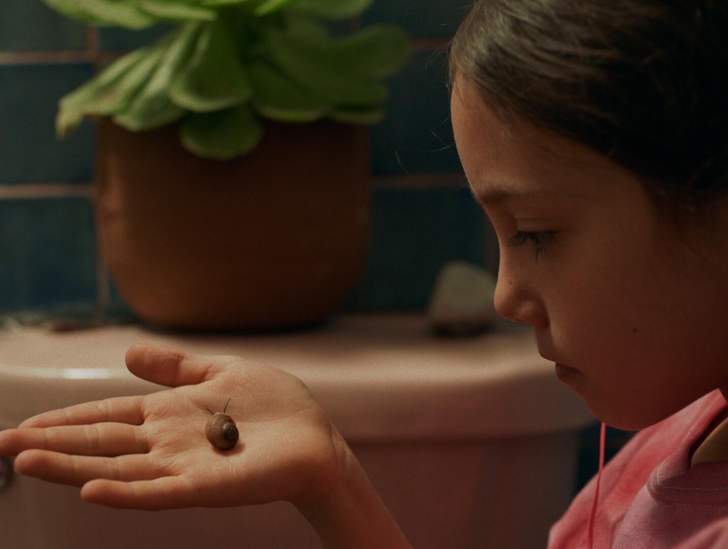 A seemingly young individual holding out their hand with a small snail on it. In the background, there is a potted plant sitting on what appears to be a bathroom sink or countertop.