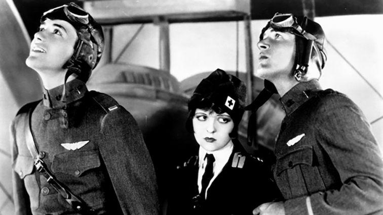 Three individuals dressed in military pilot uniforms standing in front of an aircraft. The uniforms have badges and insignia, suggesting a rank or affiliation with an air force. The setting appears historical due to the style of the uniforms and the desig