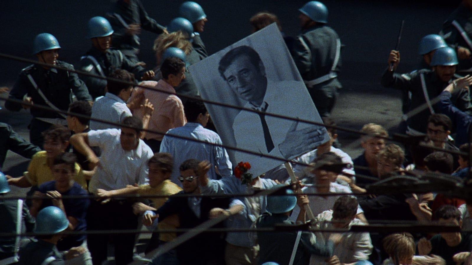 A street scene with a large crowd of people gathered, holding signs and banners. The atmosphere appears tense, suggesting a protest or demonstration. Buildings and trees line the street, and the sky is overcast.