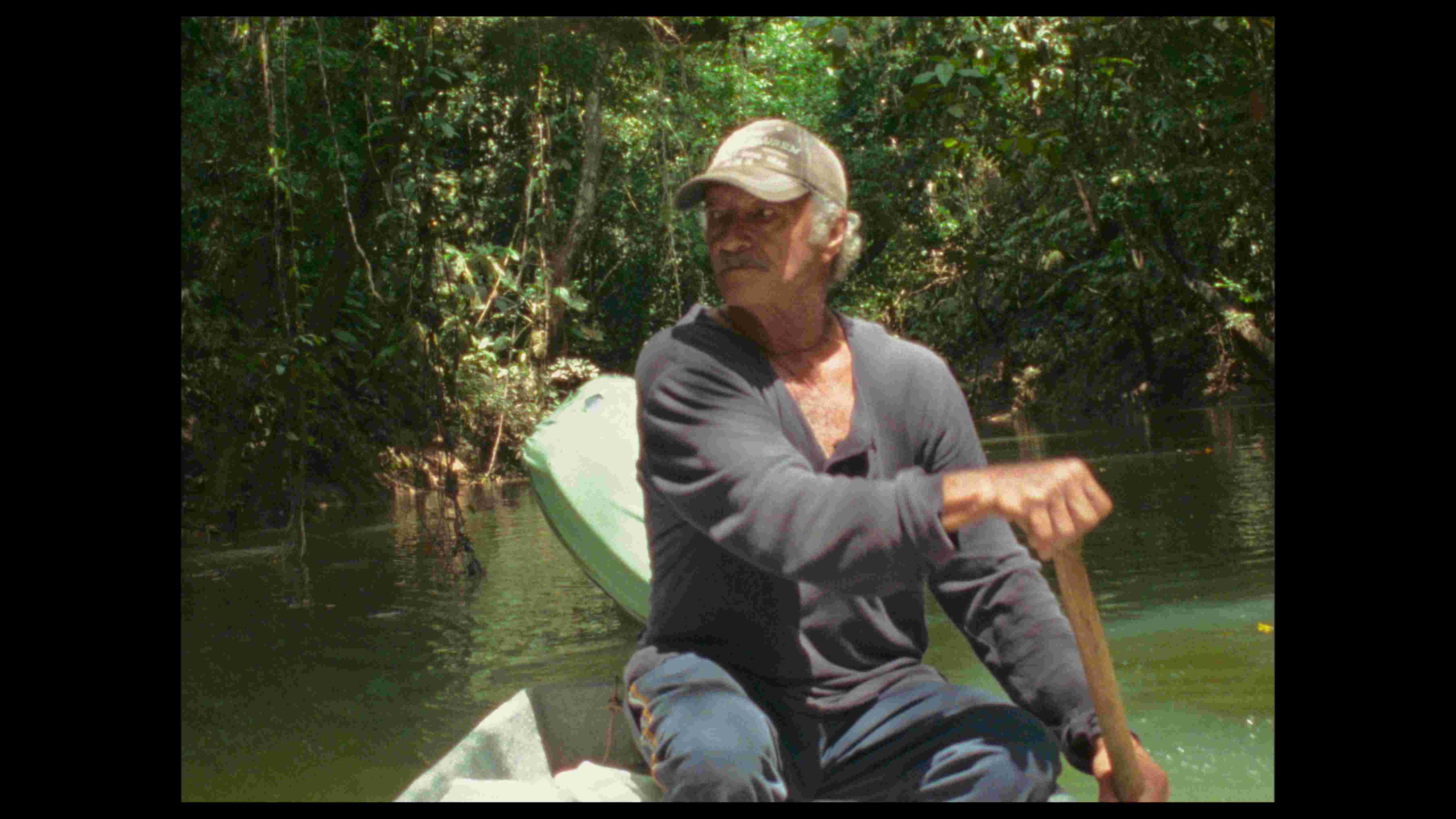 A man wearing a baseball hat rowing a boat on a lush river.