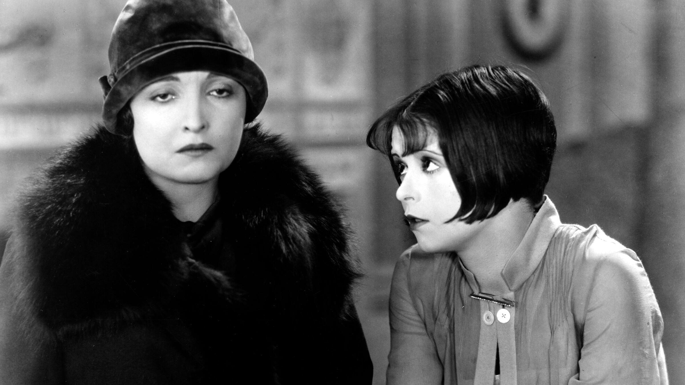 A close up shot of two women standing next to each other, one wearing a hat and one with a bobbed haircut.