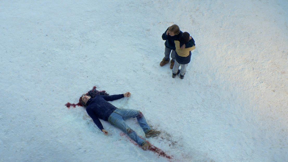 Two people standing next to a dead body in the snow with a bloody head injury.